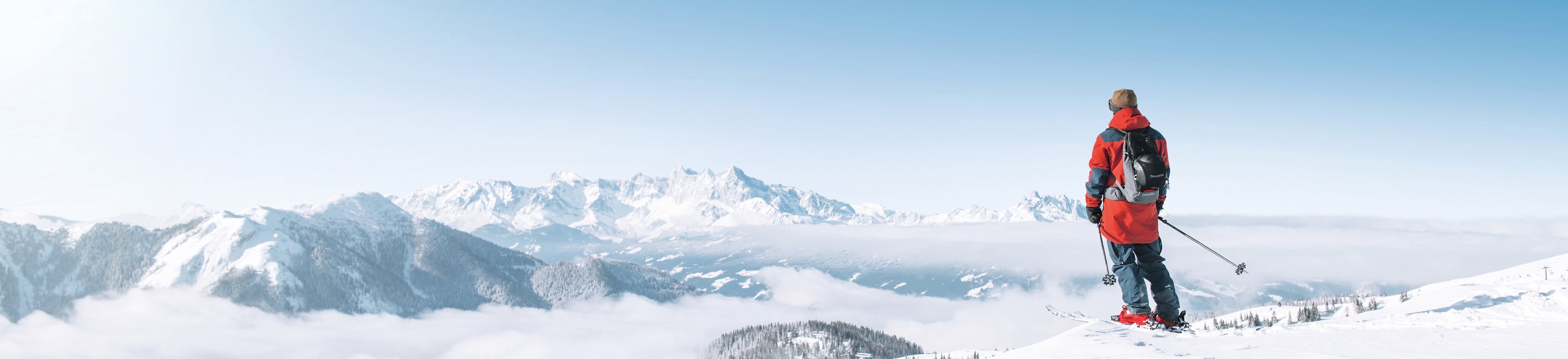 Skier overlooking snow-covered mountains, fog in the valley, and a bright blue winter sky.