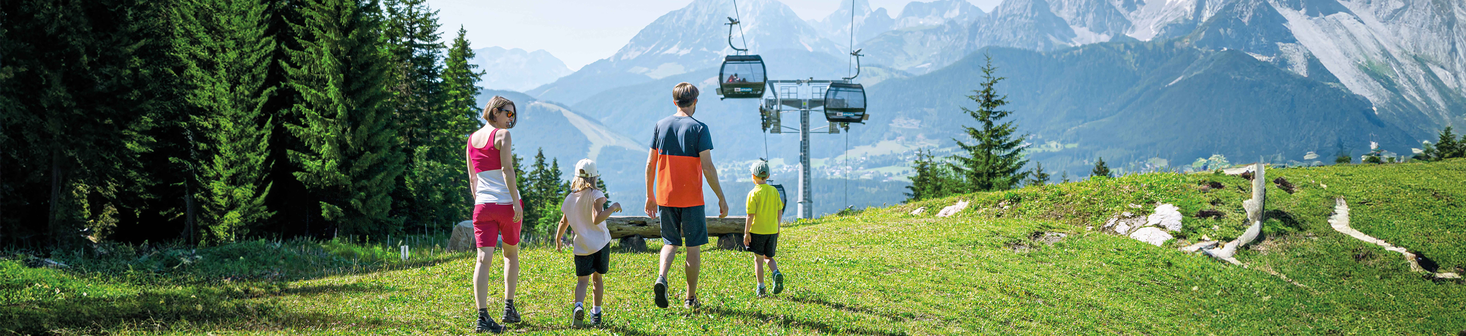 Familie wandert bei Sonnenschein durch grüne Berglandschaft, im Hintergrund fährt eine Gondel.